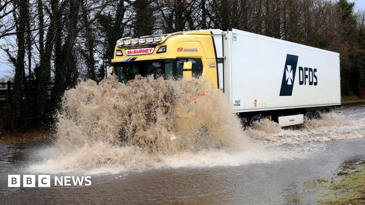 A large yellow and white lorry is driving into a flood. The water is covering the can and the road.