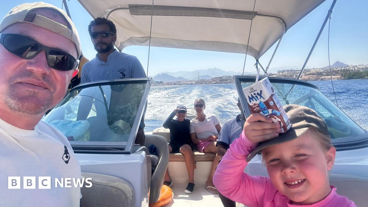 A man in sunglasses is taking a selfie on a boat with water and hills in the background, his daughter is beside him in a gap, squinting slightly and smiling, a carton of what looks like chocolate milk on her head. A boy and a woman in sunglasses are further back on the boat and a man with a T-shirt saying 'Deep Blue Dive Club' and sunglasses stands up, appearing to work on the boat.