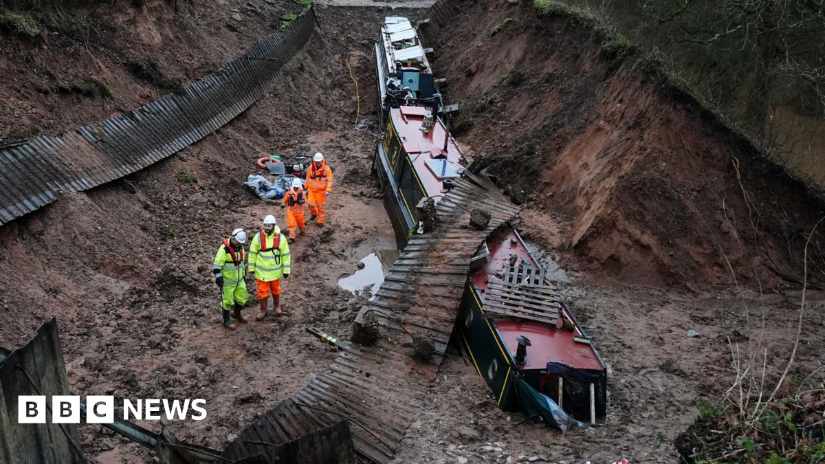 Millie Bobby Brown and David Harbour in character as Eleven and Jim Hopper. Eleven wears red shorts over grey leggings and a grey jumper. Jim Hopper wears a black leather jacket, a black beanie hat and dark trousers.They are speaking to director Frank Darabont on the set of Stranger Things. There is a vintage advertisement posted in the background with debrief on the ground.