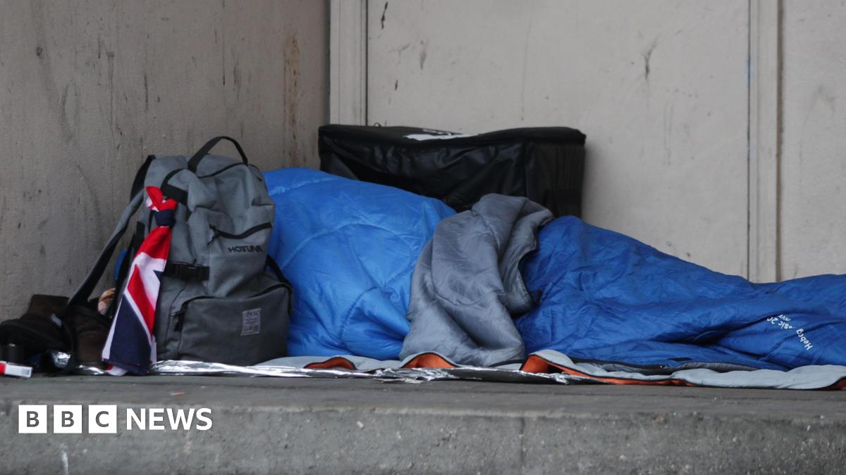 A rough sleeper in a blue sleeping bag sleeps in a doorway with a grey ruck sack by their side.