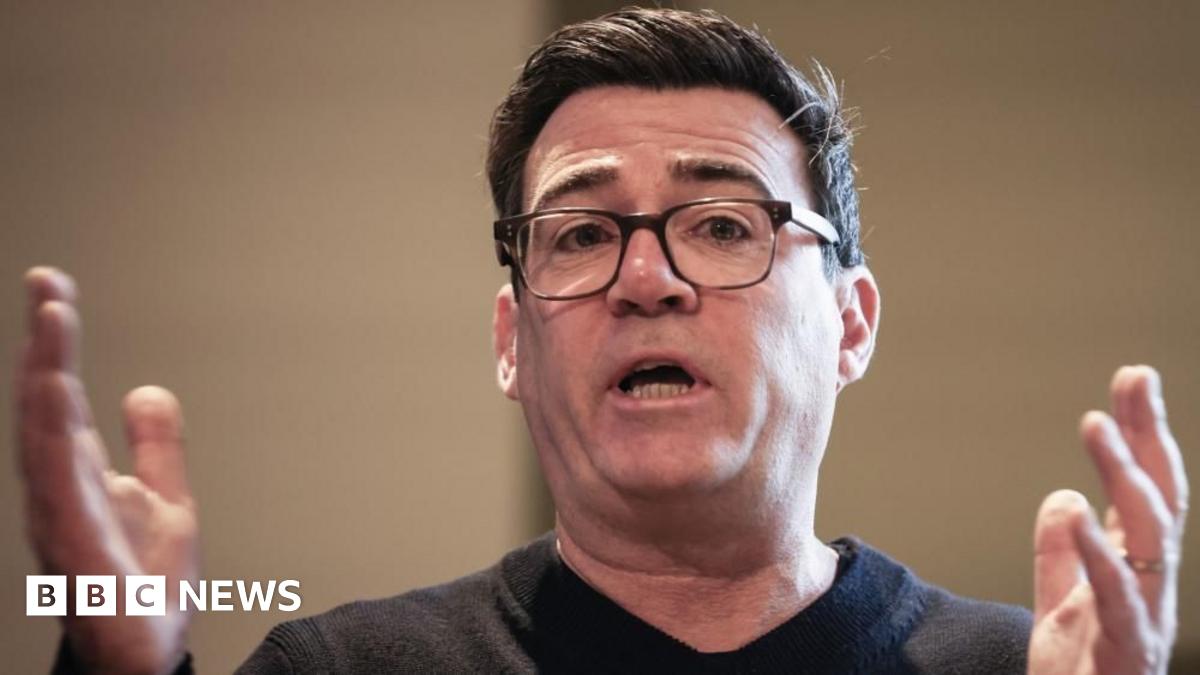 Andy Burnham is wearing a black jumper and black glasses. He is speaking with his hands raised and held apart.