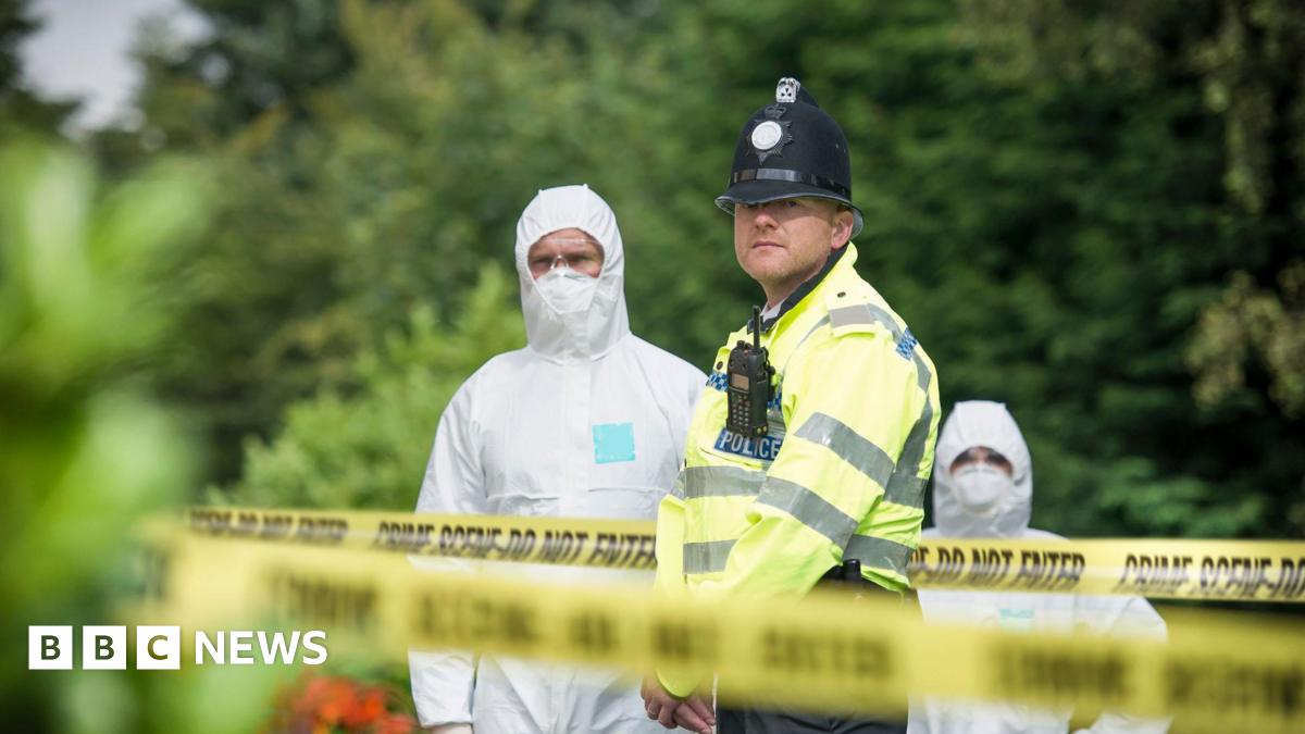 Police officer standing by crime scene tape with forensic investigators in white overalls and trees behind him.