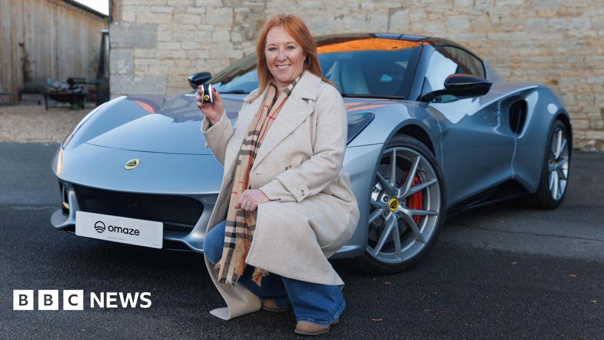 Claire Carty is crouched in front of a grey Lotus car. She has mid length red hair and is wearing a beige jacket and a checked scarf and brown boots and is holding up a set of car keys and smiling at the camera.