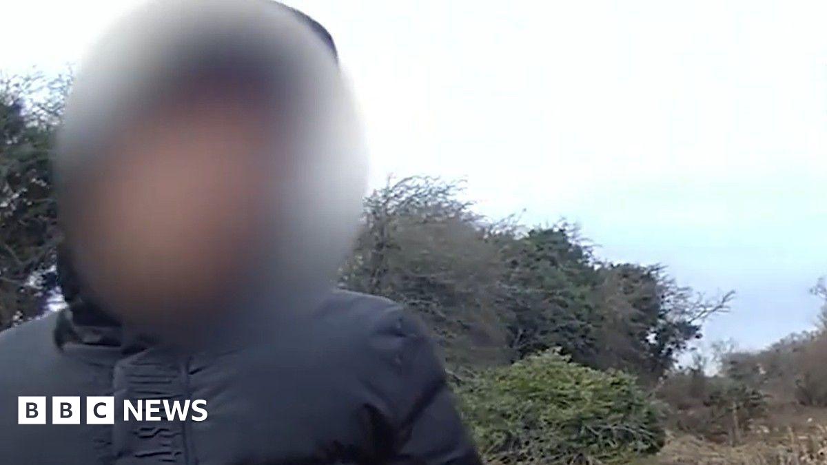 A man with a bald head in a blue suit with a red tie in a room with white walls and a blurred blue symbol on one wall