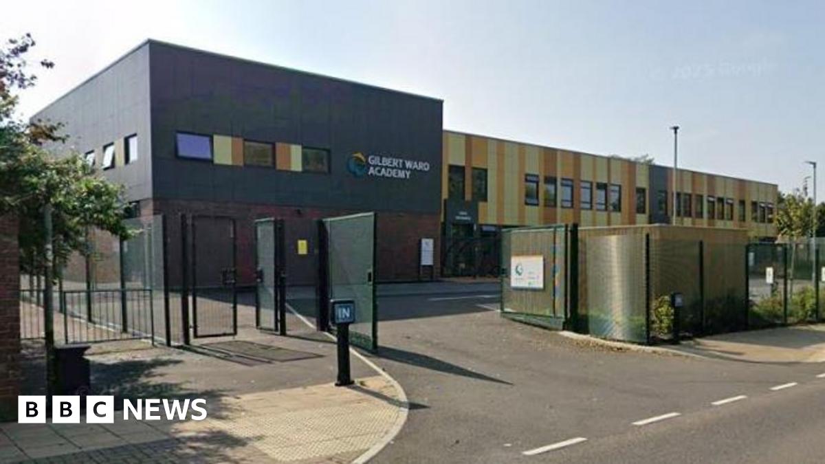 Gilbert Ward Academy, a modern school building with brown and yellow cladding. There is a tarmac entrance and a metal fence.