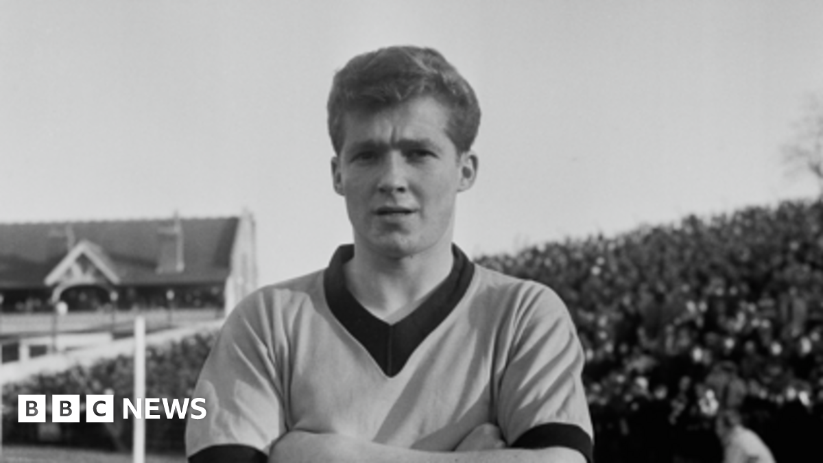 A black and white image of footballer Terry Wharton at a game in 1961. The photo is a close-up of Wharton, who is crossing his arms across his chest, while standing on a football pitch. The crowd is visible behind him.