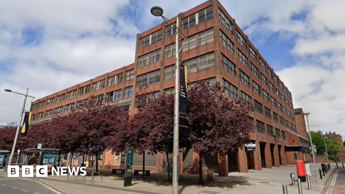 A Google Streetview screenshot of Vancouver House in Middlesbrough. The large six-storey office block  is built in red brick with large rectangular windows on each floor. The building gives onto a main road where there is a bus stop.