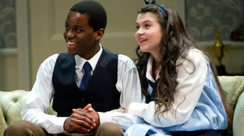 RCS Two people sitting closely on a light-colored sofa in a staged setting. One is wearing a white shirt with a dark vest and tie, while the other is dressed in a light blue outfit with a matching headband. Their hands are clasped together, and the background includes decorative elements such as a trophy and patterned walls.