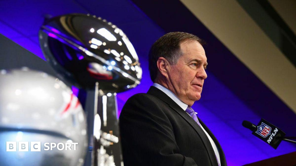 Bill Belichick, pictured in 2019, talking into an NFL-branded microphone at a press event. He has short dark hair and is wearing a dark suit with a pale short and purple tie. To his right are the Super Bowl trophy and a New England Patriots helmet.