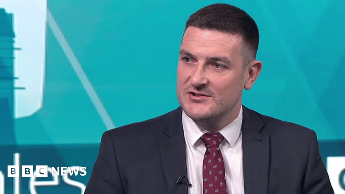 A man with short brown hair and a navy suit and red tie speaks on camera.