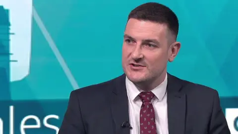 BBC A man with short brown hair and a navy suit and red tie speaks on camera.