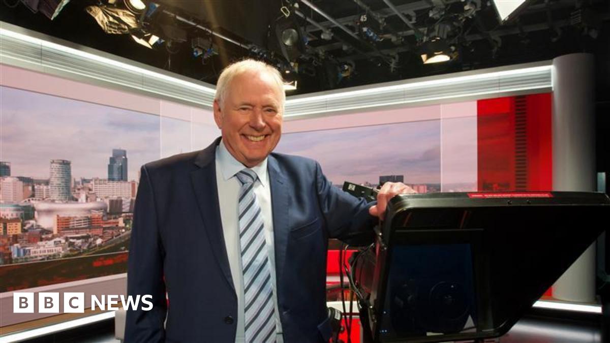 BBC TV presenter Nick Owen stands in the studio leaning on one of the cameras, while smiling. The Birmingham landscape is projected on the screen behind him.