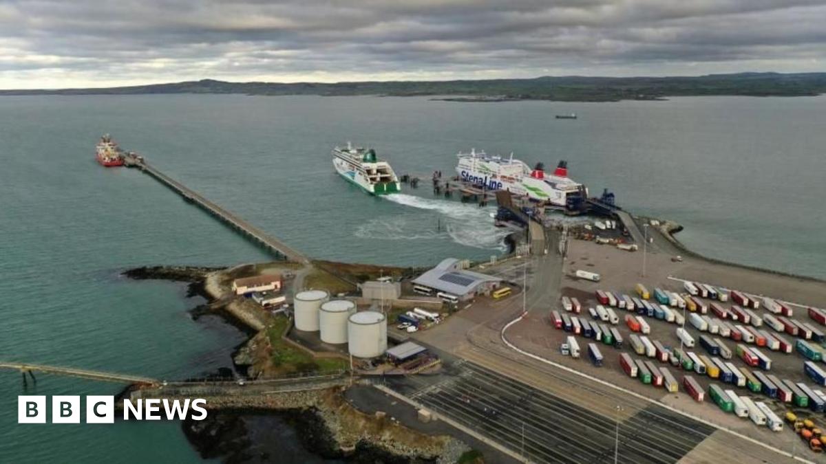 A concrete dock, with two boats in the sea in front of it, and a car park to one side, full of vehicles, with various industrial buildings on the other side of the dock