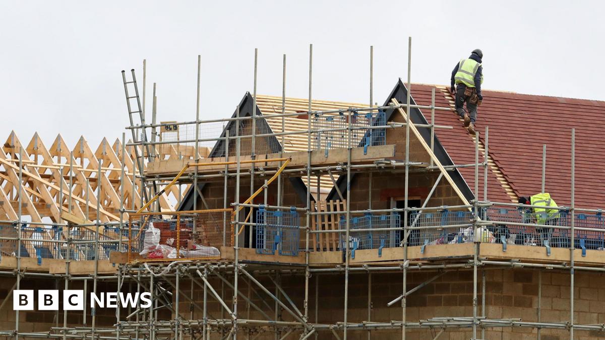 Two workers wearing hi-vis jackets work on the roof of a new, partially built house on a construction site.
