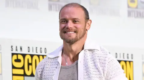 Getty Images Heughan wearing a light gray tank top under a white short-sleeve crochet shirt, standing in front of a backdrop with the San Diego Comic-Con International logo in black and yellow.