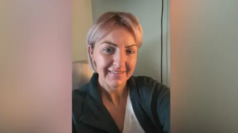 Emma Metcalfe A woman wearing a black jacket and grey t shirt is taking a selfie. Behind her is a wire and a blue and white wall