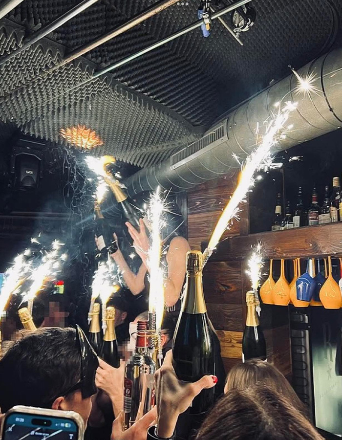 Crowd celebrating with champagne and sparklers in a lively bar scene related to Swiss ski resort fire allegations. Crowd celebrating with champagne and sparklers in a lively bar scene related to Swiss ski resort fire allegations.