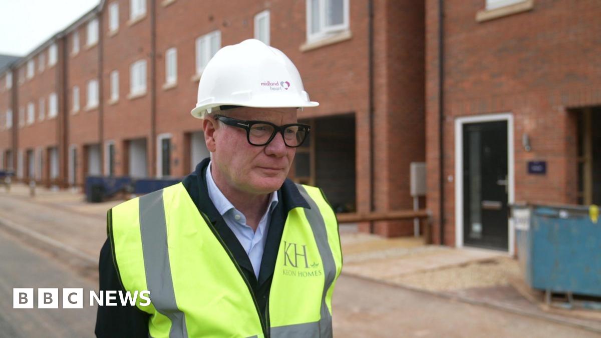 Richard Parker wearing a white builder's hat, glasses and a hi-vis jacket over a suit, stands and looks to our right. Behind him is a row of homes that are under construction.