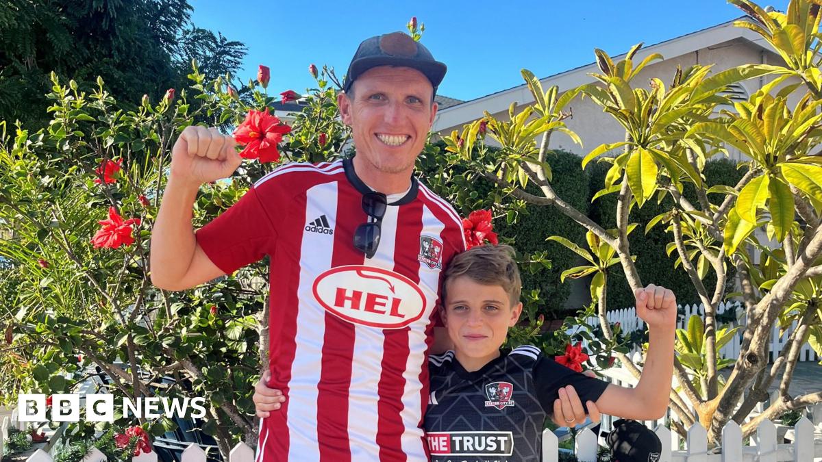 George and Charlie standing together. They are both holding one arm up in the strong position and wearing Exeter City shirts. George is wearing a white and red and top while Charlie is wearing a black and white shirt. There are plants and trees behind them and it is a sunny day.