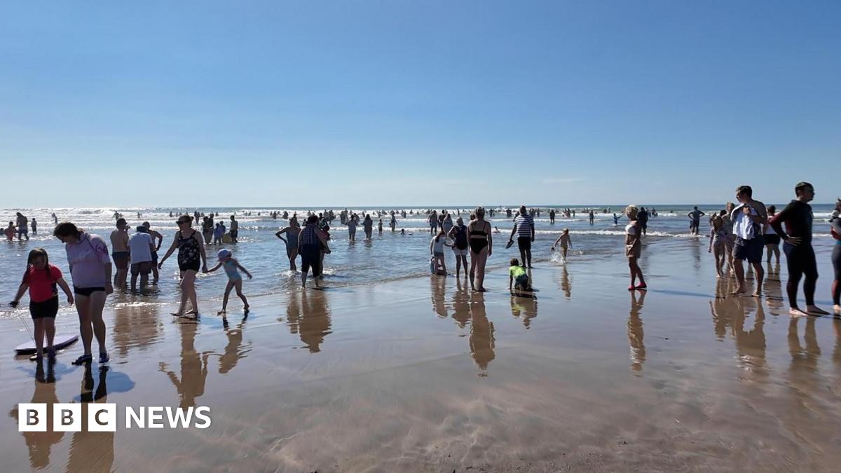A view of a beach, with swimmers and people with body boards standing on the sand. In the distance you can see people in the water