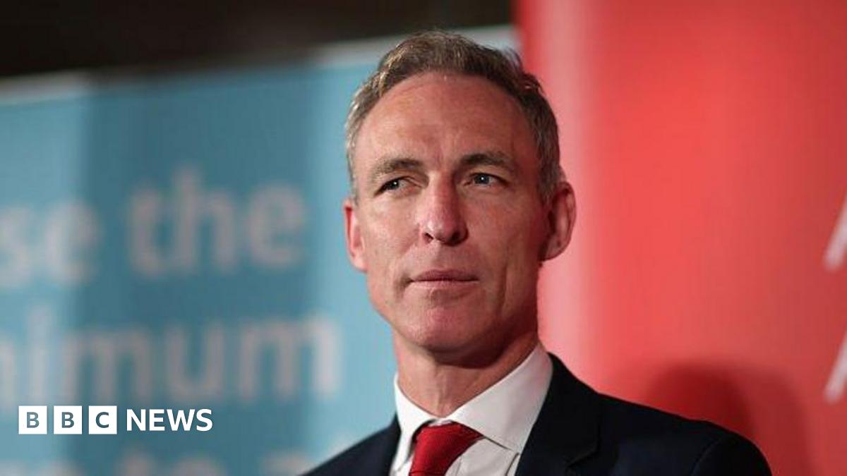 Jim Murphy wearing a neutral expression in a dark suit and red tie against red and blue background at a Labour Party event in 2015