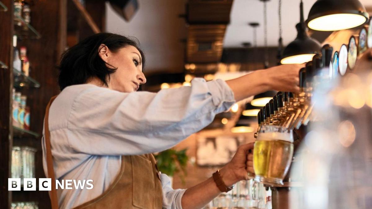 Woman with  black, shoulder-length hair, wearing a light blue shirt and brown apron pulls a pint of lager behind the bar in a pub