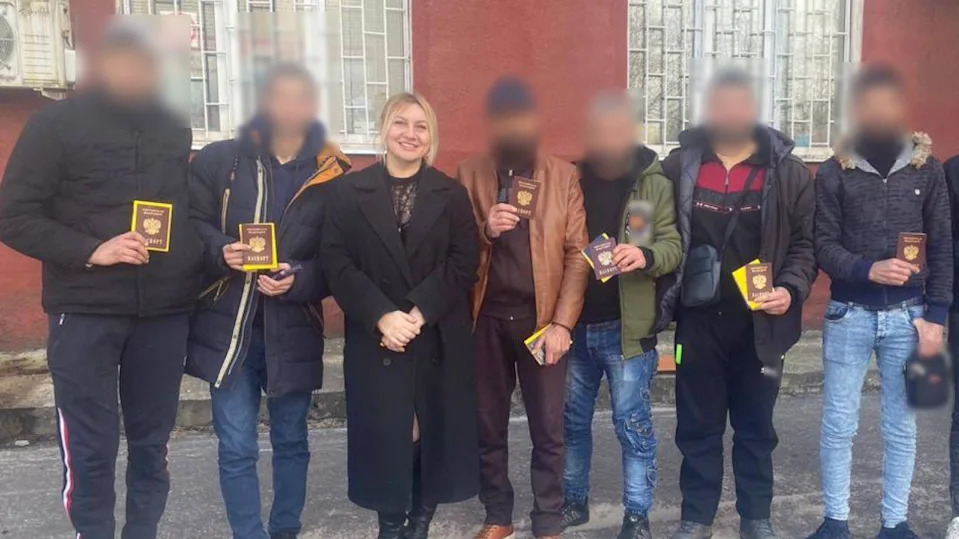 Polina Azarnykh standing smiling in a long black coat and smart black boots in a line of men wearing jeans, casual shoes and jackets. Their faces have been blurred to conceal their identities. They are holding up Russian travel documents.