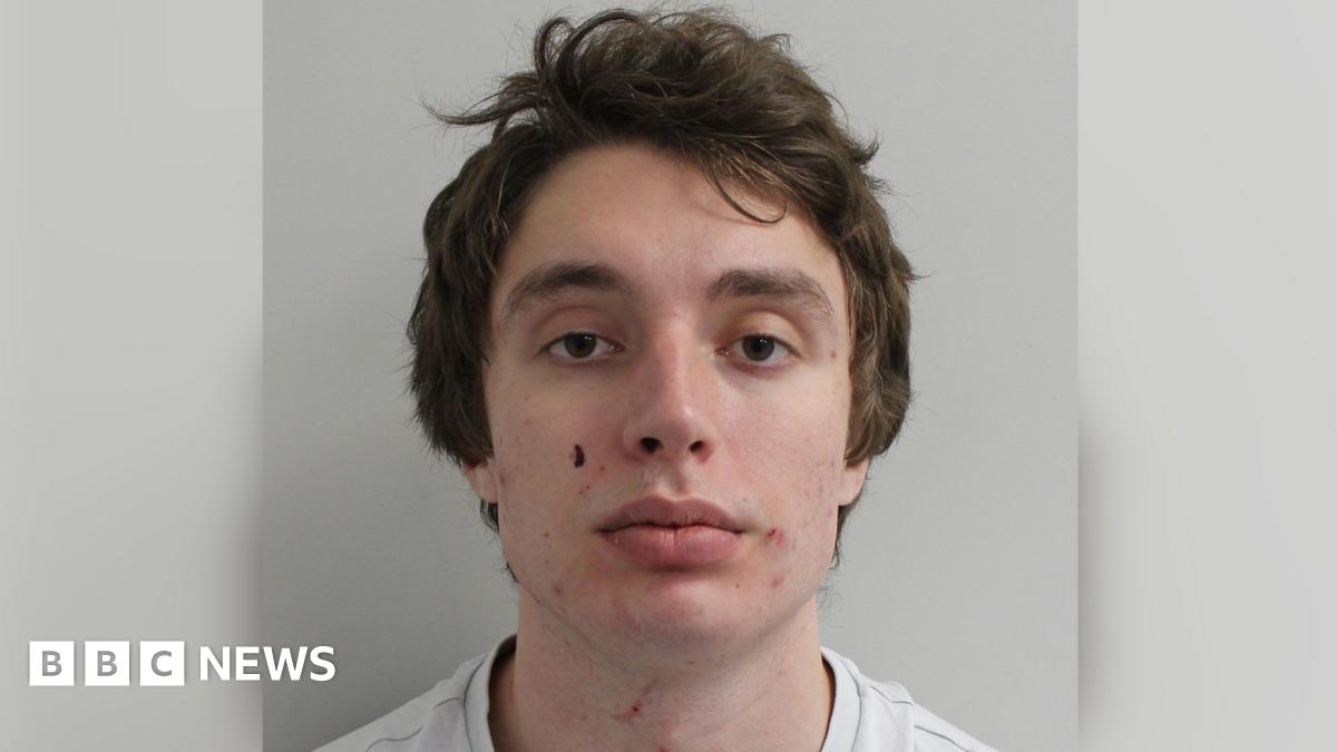 Custody image of Matvei Rumiantsev. He is a young man with short brown hair, wearing a white t-shirt. He stands with a neutral expression in front of a white wall.