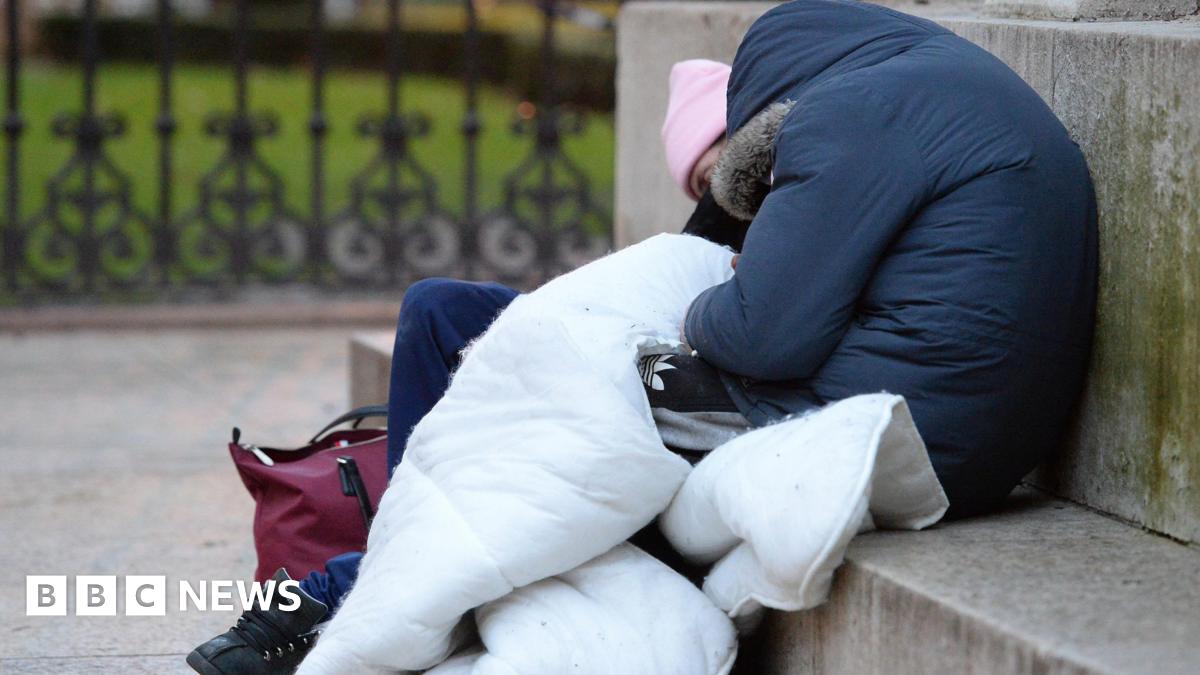 A person sat slumped over on a concrete step with their blue hooded coat covering their face as they sleep. They are holding a white duvet  while sleeping next to someone else who wears a pink beanie hat, blue jeams and black nike trainers. There is a maroon handbag on the floor next to them
