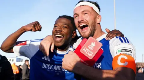 PA Media Macclesfield FC goal scorers Isaac Buckley-Ricketts and Paul Dawson celebrate following the Emirates FA Cup third round match at the Leasing.com Stadium, Macclesfield. They are wearing Macclesfield FC kits and have big smiles, with Buckley-Ricketts raising his arm in the air in celebration. Dawson, with a captain armband, holds a card with the Emirates FA Cup logo on it.
