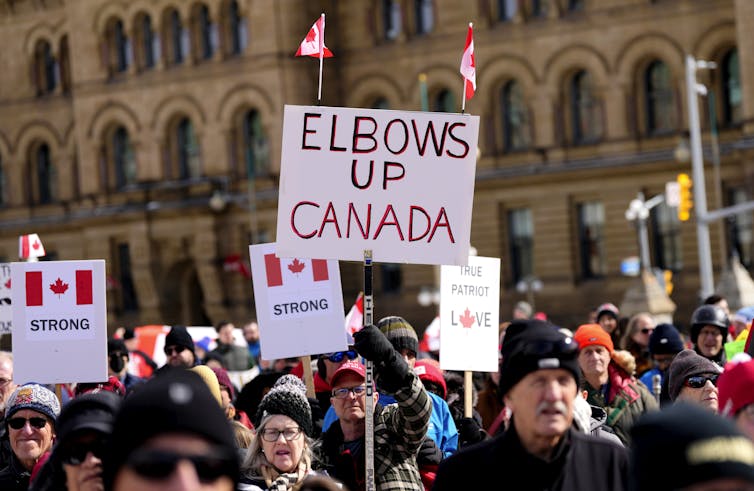 a man at a demonstration carries a placard with a sign reading: elbows up Canada