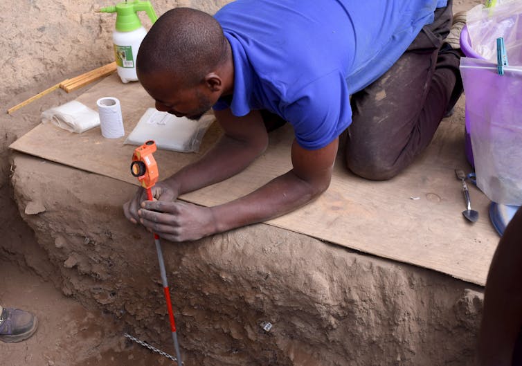 man kneeling on a board measures down into the excavated area
