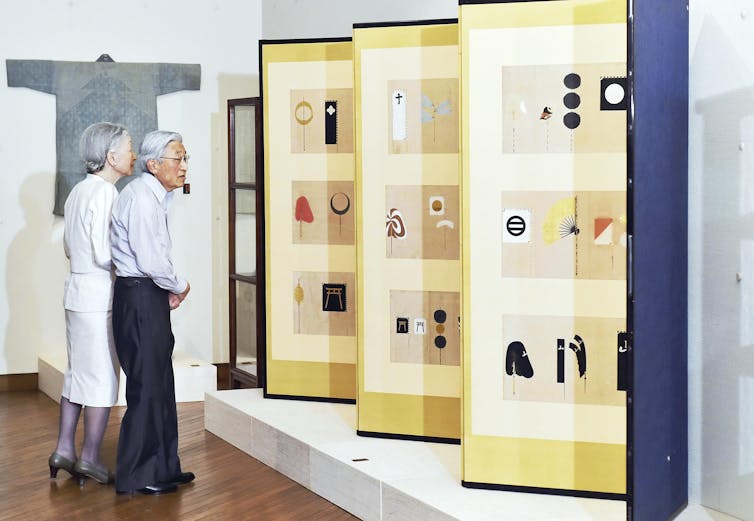 An older Japanese man and woman look at some folk art on panels inside a gallery.