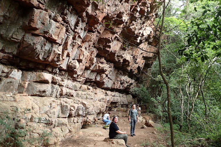 Woman sitting under a rockface, man standing in background.