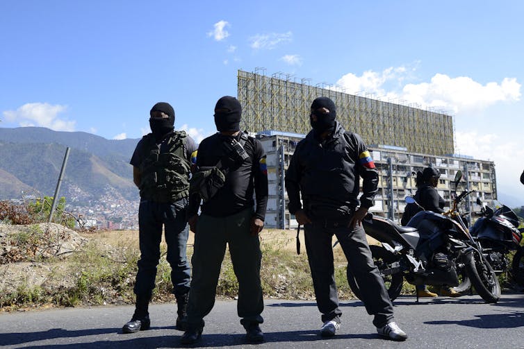Three masked and armed men stand guard.