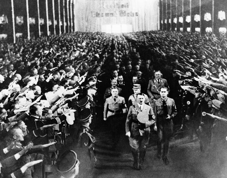 A black-and-white photo shows uniformed soldiers saluting Adolf Hitler as he walks into a gathering.