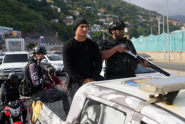 Three armed police officers ride in the back of a pickup truck.