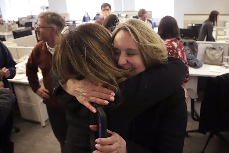 Two women hug in foreground while people stand around desks in background