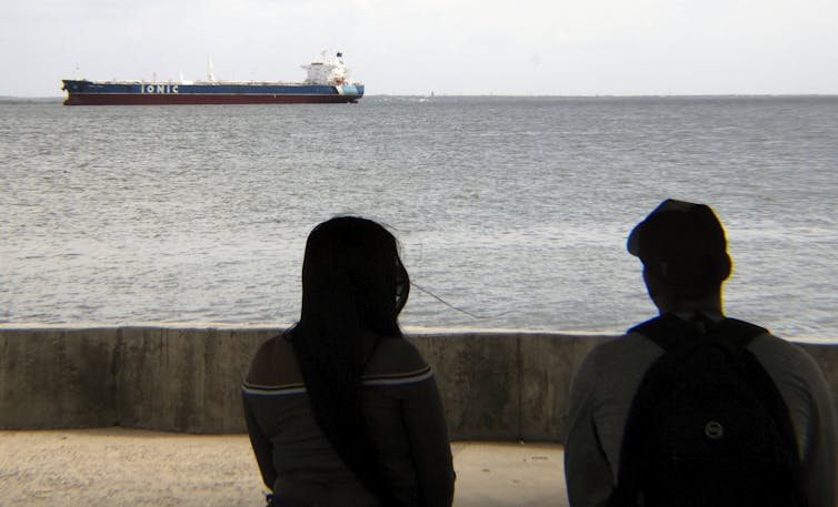 Two figures watch a large ship move across the water.