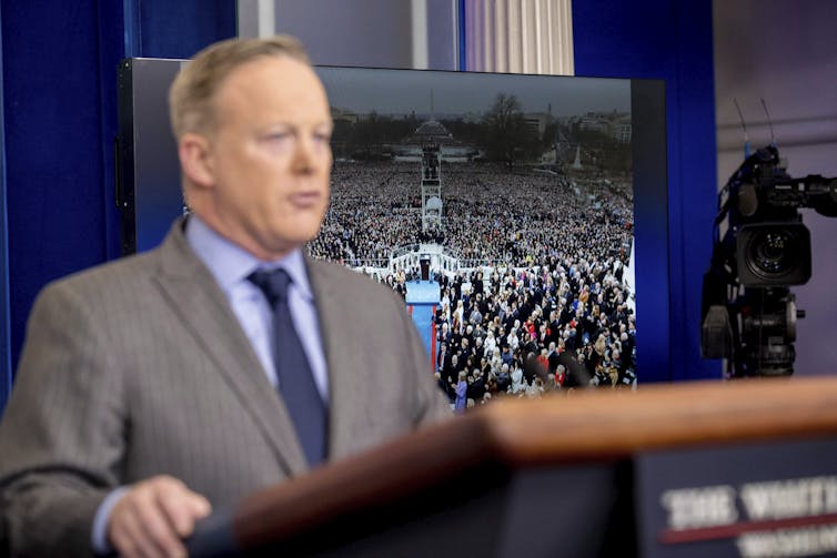 A man behind a podium looks nervous. A photo of a crowd is behind him.