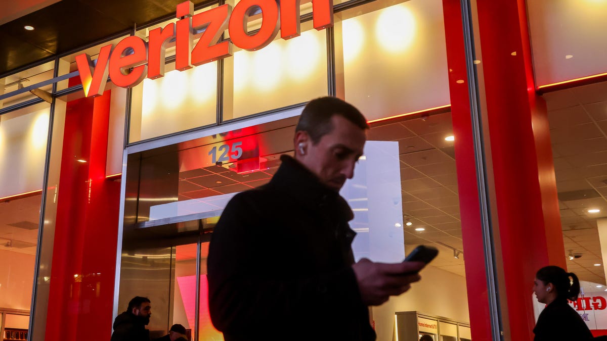 A man looking at a phone walks past a Verizon store.