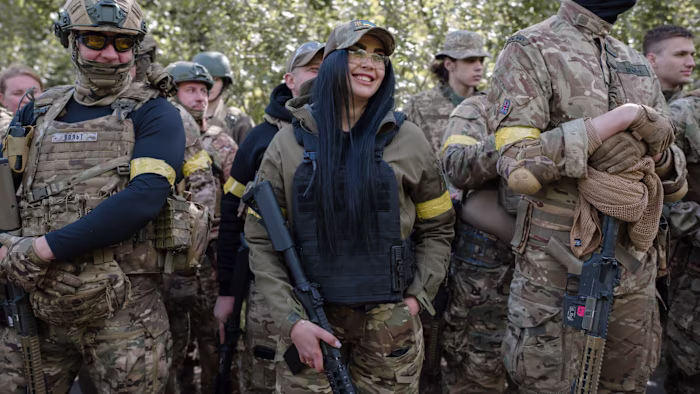 People in camouflage uniforms with yellow armbands stand together, some holding rifles