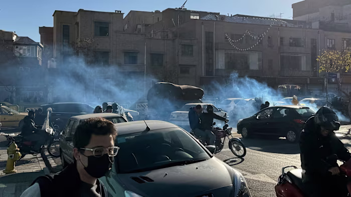 Tear gas fills a street as people, some on motorcycles and wearing masks, move among cars during a protest in Tehran.
