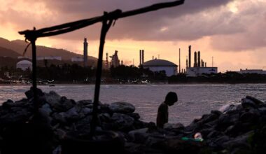 A child walks along a rocky beach at dusk, with the El Palito refinery's industrial structures silhouetted in the background.