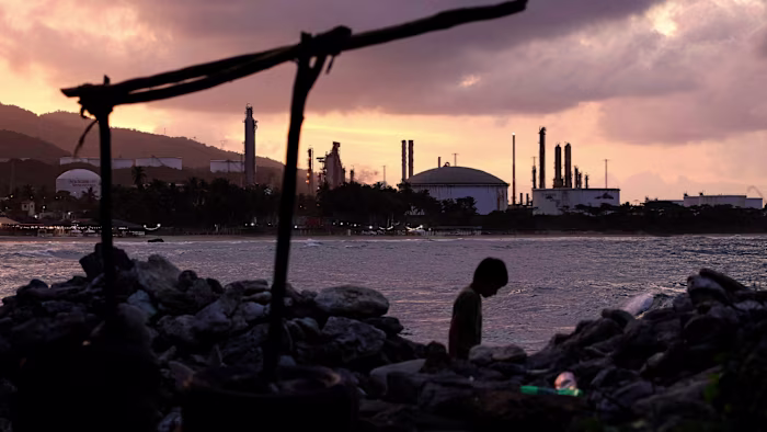 A child walks along a rocky beach at dusk, with the El Palito refinery's industrial structures silhouetted in the background.