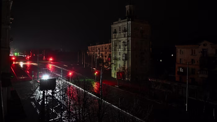 A near empty city street in Zaporizhzhia, illuminated only by car headlights and faint traffic lights during a blackout.