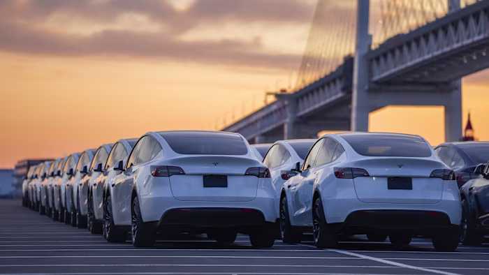 Rows of white Tesla vehicles parked in a lot near a bridge at sunset, newly arrived at a port in Yokohama, Japan.