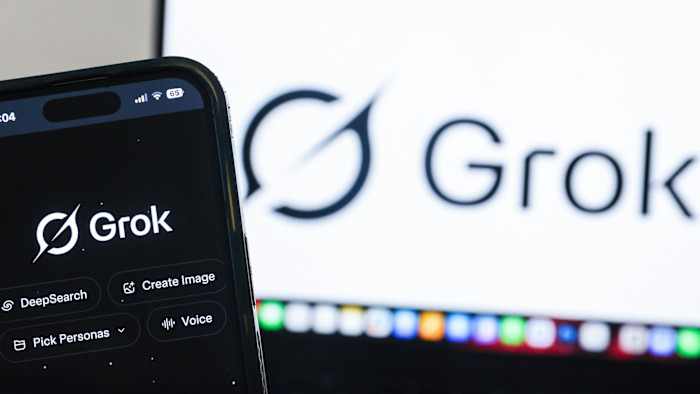 The Grok AI website is shown on a smartphone screen with the Grok logo also displayed on a computer monitor in the background.