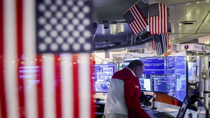 A trader in a red jacket works at a desk with multiple monitors on the New York Stock Exchange floor, with several American flags visible.