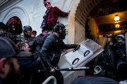 a crowd of Trump supporters in helmets and gas masks presses into a boarded-up arched entrance of the U.S. Capitol in Washington, D.C., as one swings a baseball bat and another carries a clear riot shield toward police blocking the hallway.